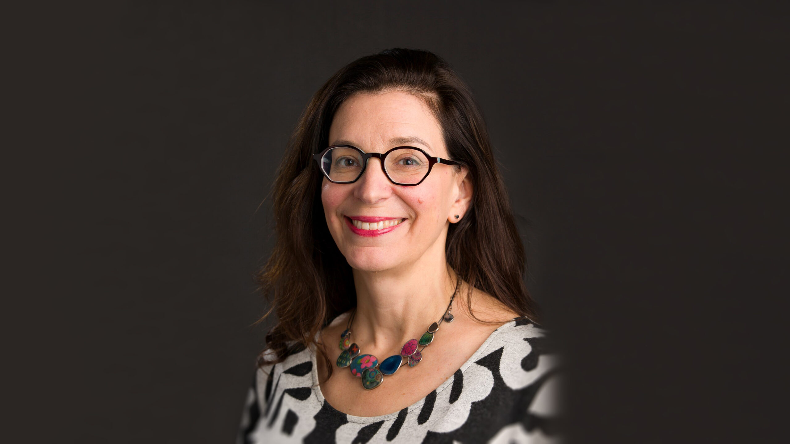 Professional headshot of a woman with dark hair and glasses wearing a colorful necklace and black and white blouse.