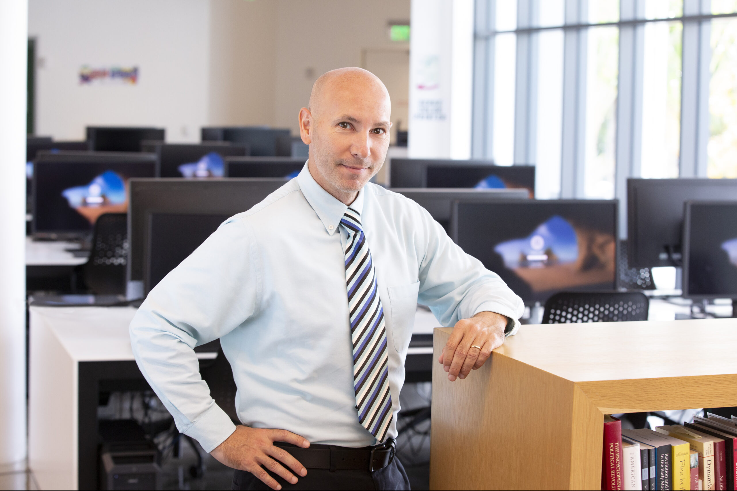 Professional headshot of a man with a striped tie, leaning on a bookshelf in a computer lab.
